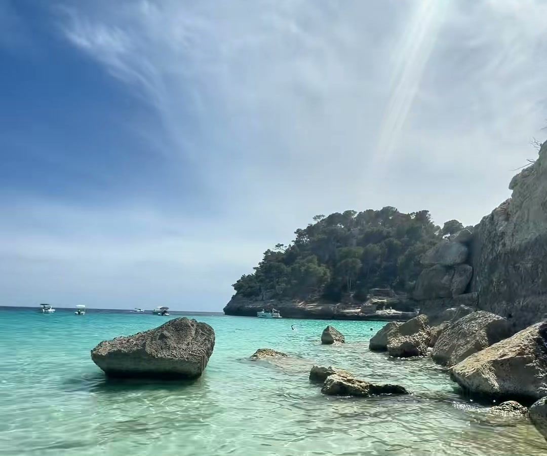 Agua cristalina y rocas en la orilla de Cala Mitjana, con el acantilado y el pinar al fondo